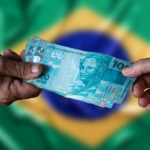 Mãos trocando notas de 100 reais com a bandeira do Brasil ao fundo.