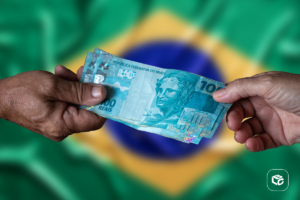 Mãos trocando notas de 100 reais com a bandeira do Brasil ao fundo.